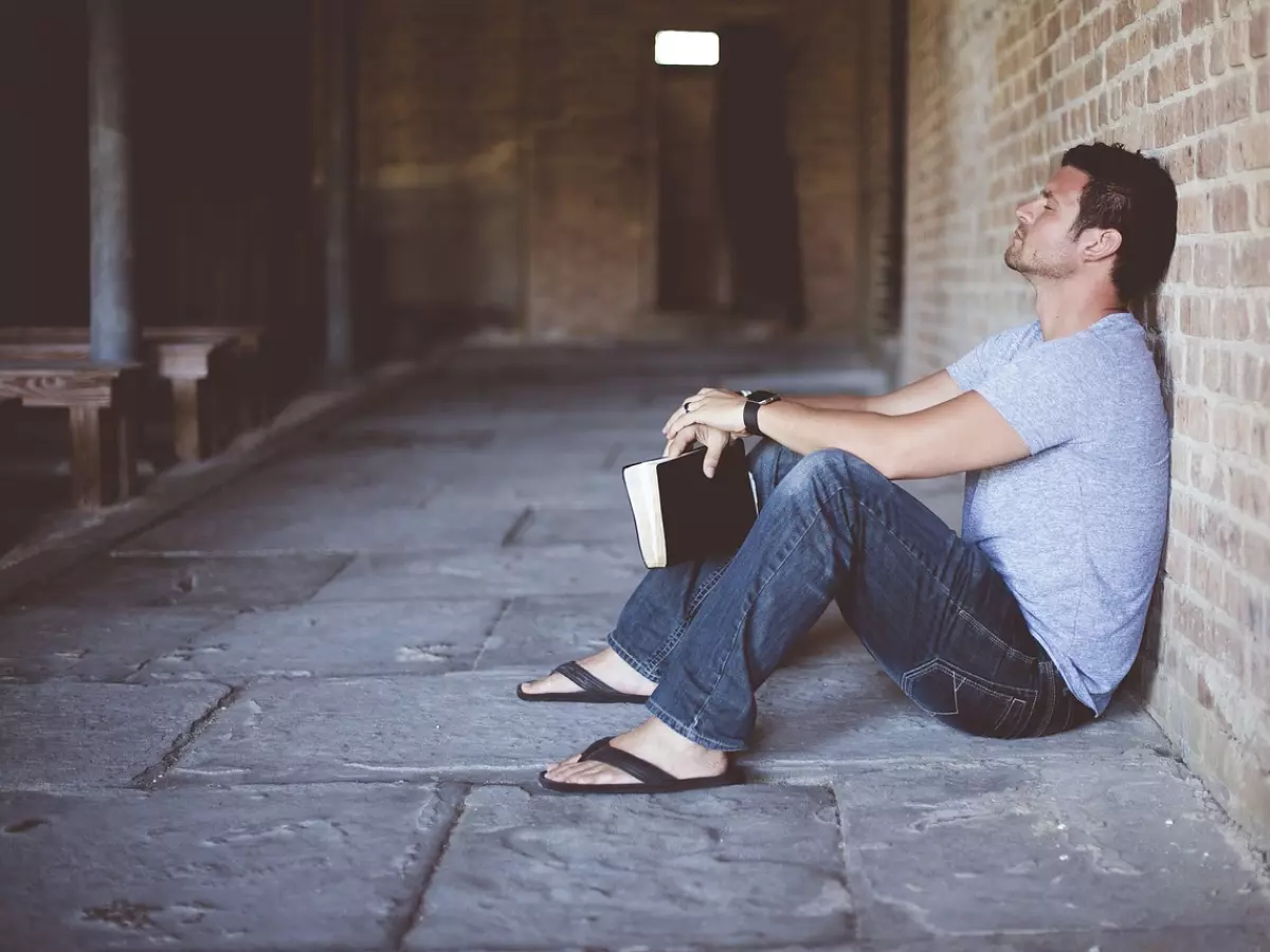 A man sitting on the floor with his back against a brick wall, holding a book in his lap. He is wearing a gray shirt and blue jeans. The photo is taken from a low angle. 