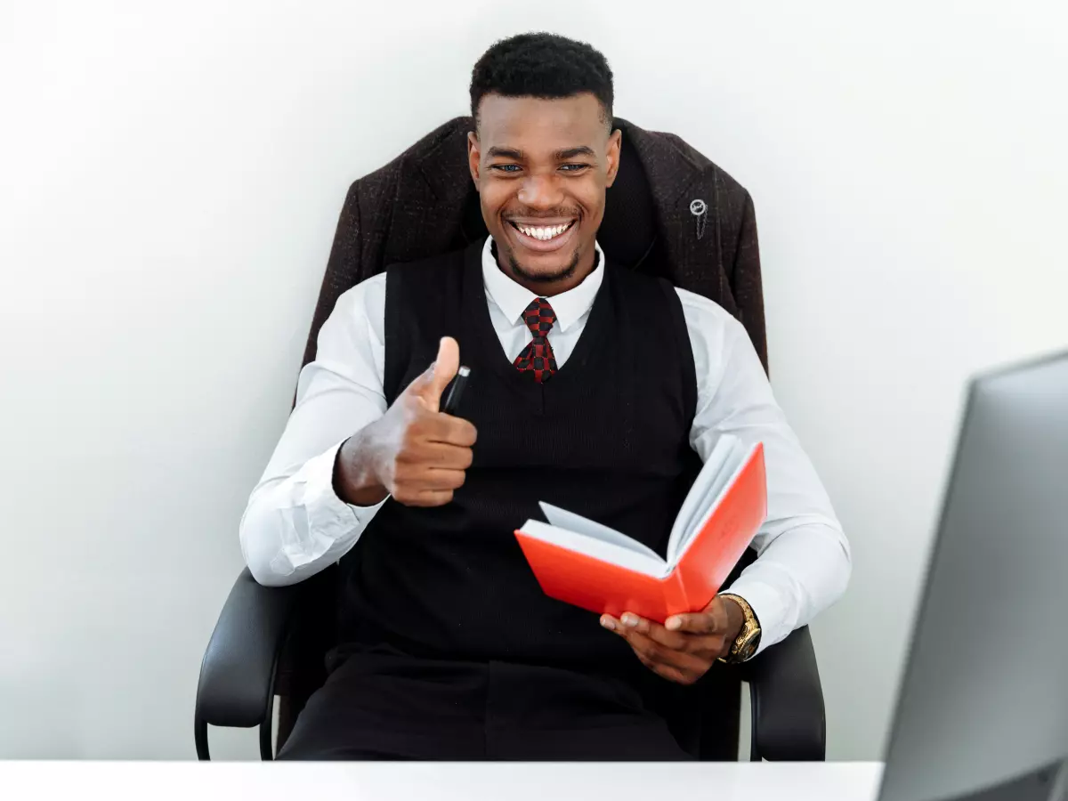 A man with a thumbs up gesture sitting in a chair, a computer monitor behind him.