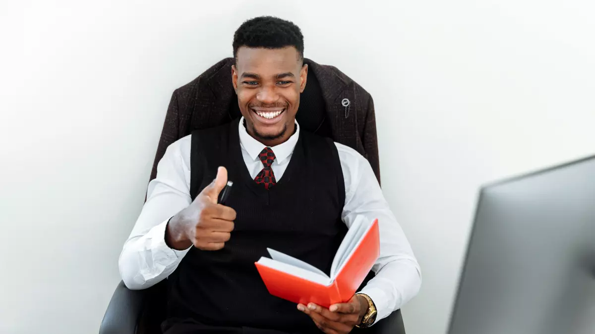 A man with a thumbs up gesture sitting in a chair, a computer monitor behind him.