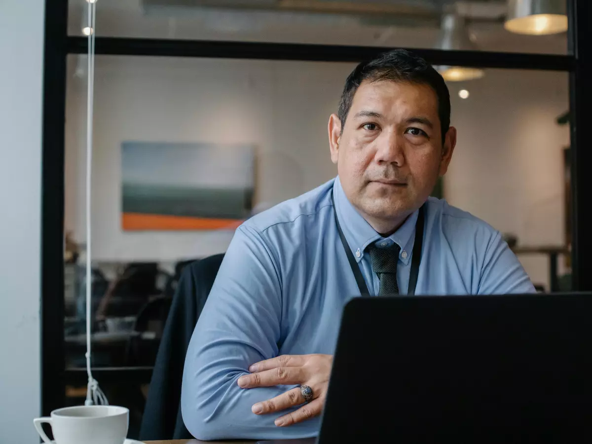 A businessman in a blue shirt and tie sits at a desk with his arms crossed and looks directly at the camera.  He is seated in front of an open laptop,  a cup of coffee on the desk.