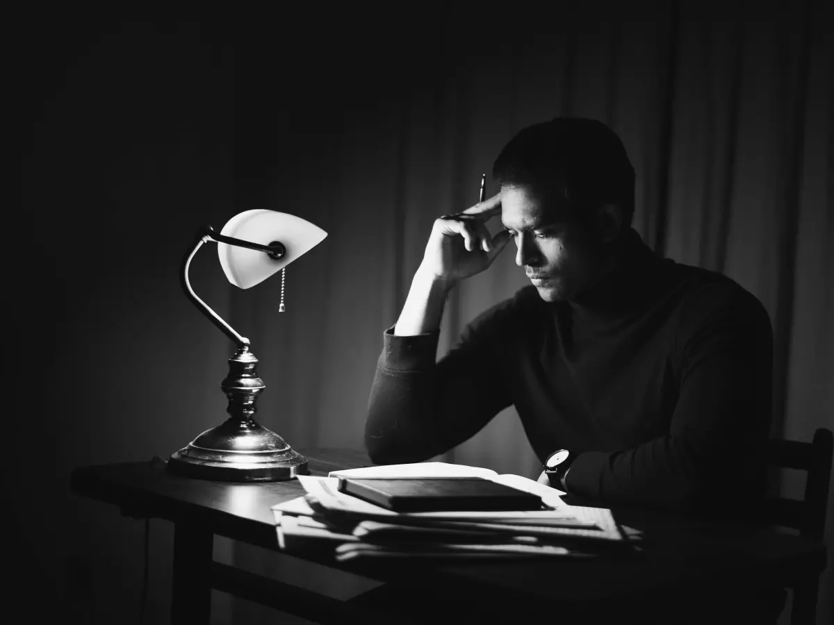 A person is reading a book in a dimly lit room. A table lamp illuminates the book and the person's face. The person is wearing a dark sweater and has their head resting on their hand.