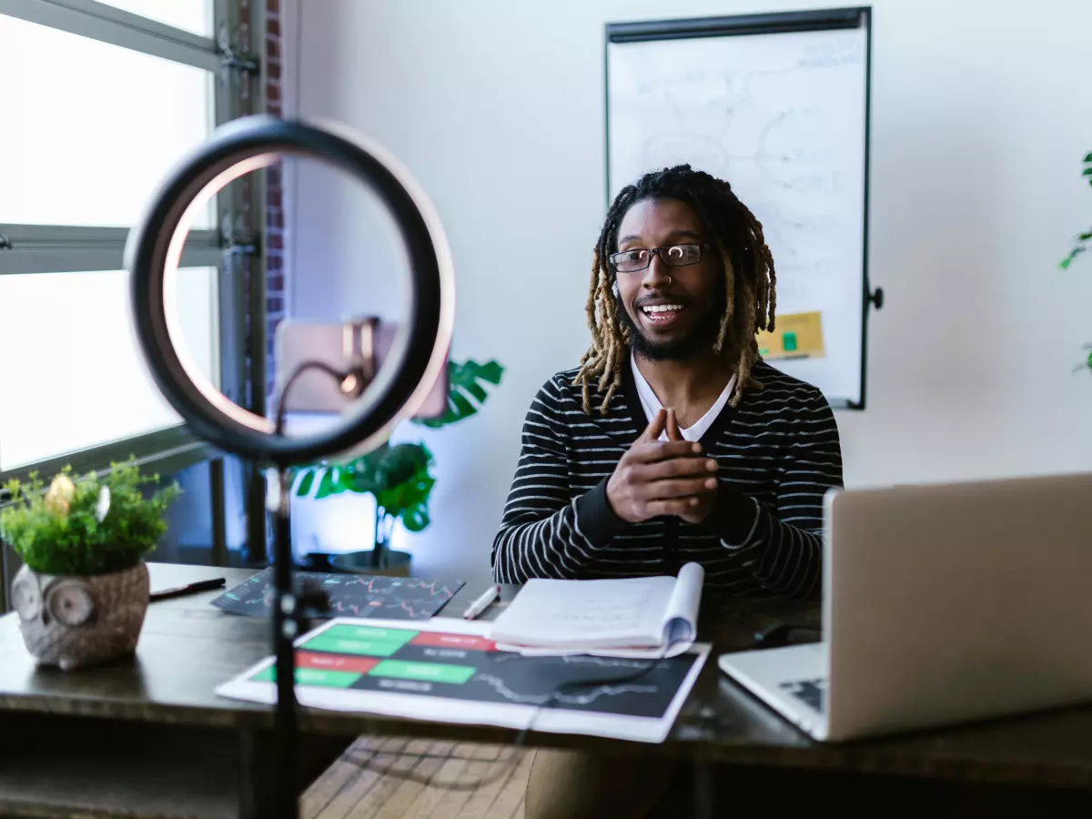 A man with dreadlocks sits at a desk in front of a laptop. He has a ring light behind him. He is smiling and looks to be happy and relaxed.