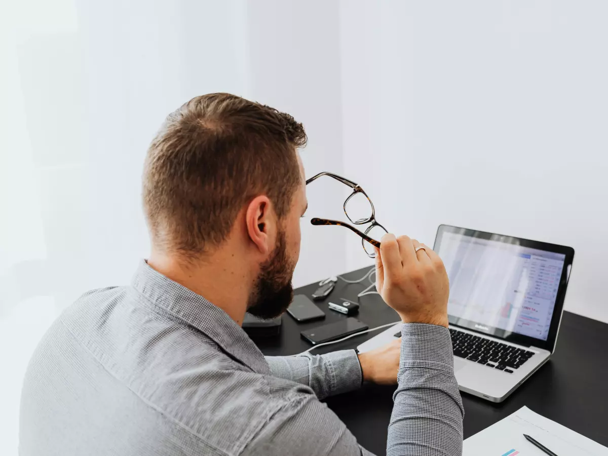 A man wearing glasses sits at a desk looking at a laptop screen in front of him.