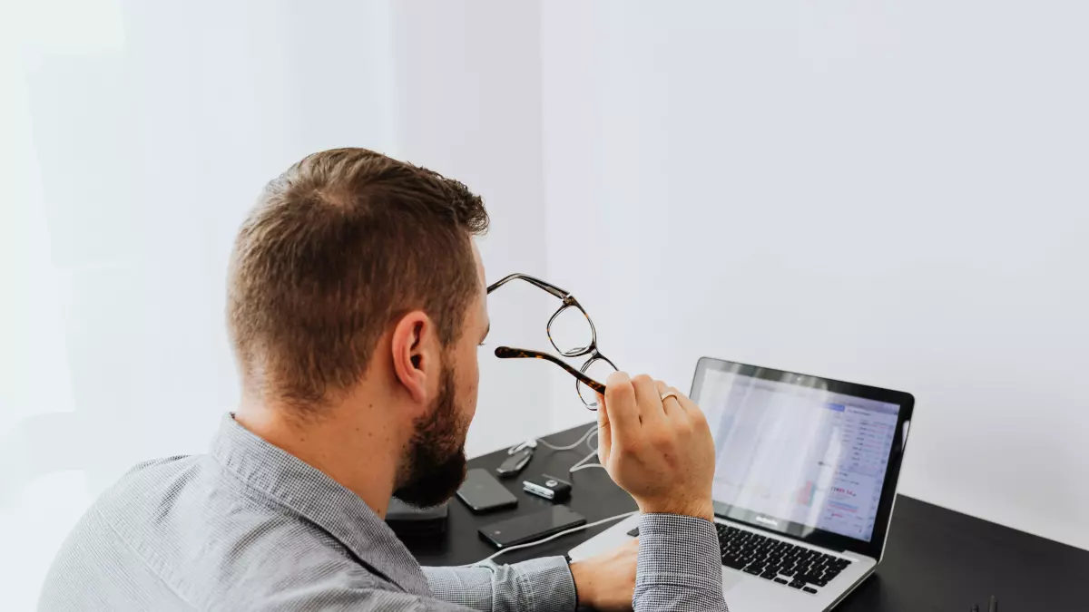 A man wearing glasses sits at a desk looking at a laptop screen in front of him.