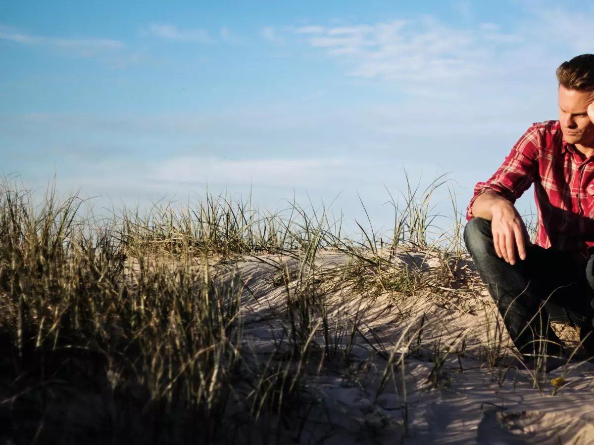 A man is sitting on a sand dune, looking down at the ground.