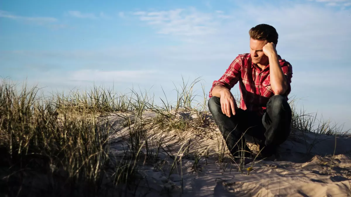 A man is sitting on a sand dune, looking down at the ground.