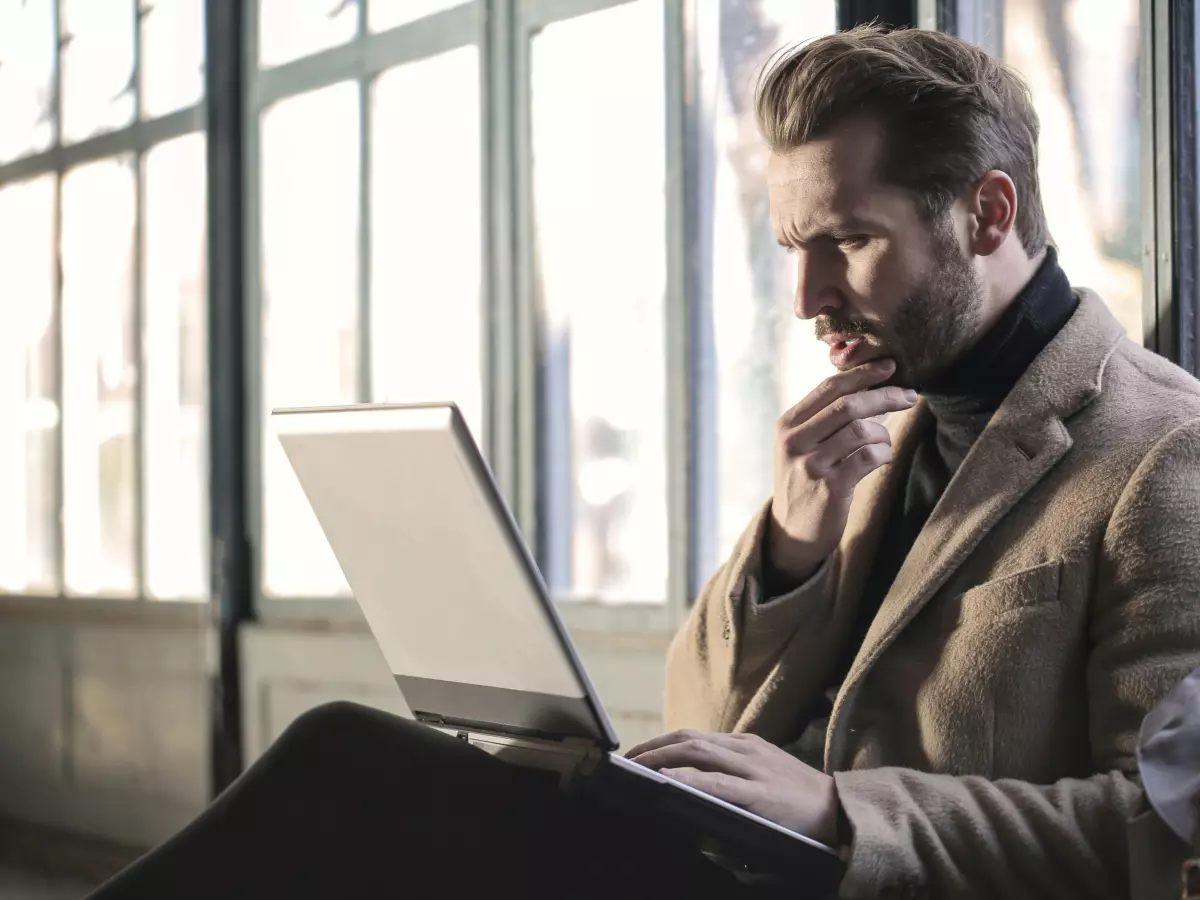 A man sitting on the floor with his legs crossed, looking at a laptop with a confused expression.