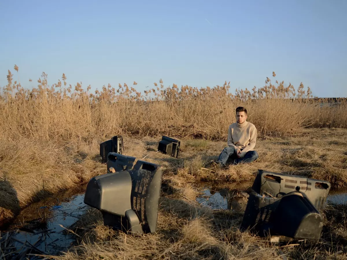 A man is sitting in a field with a bunch of old TVs around him, looking at the camera.