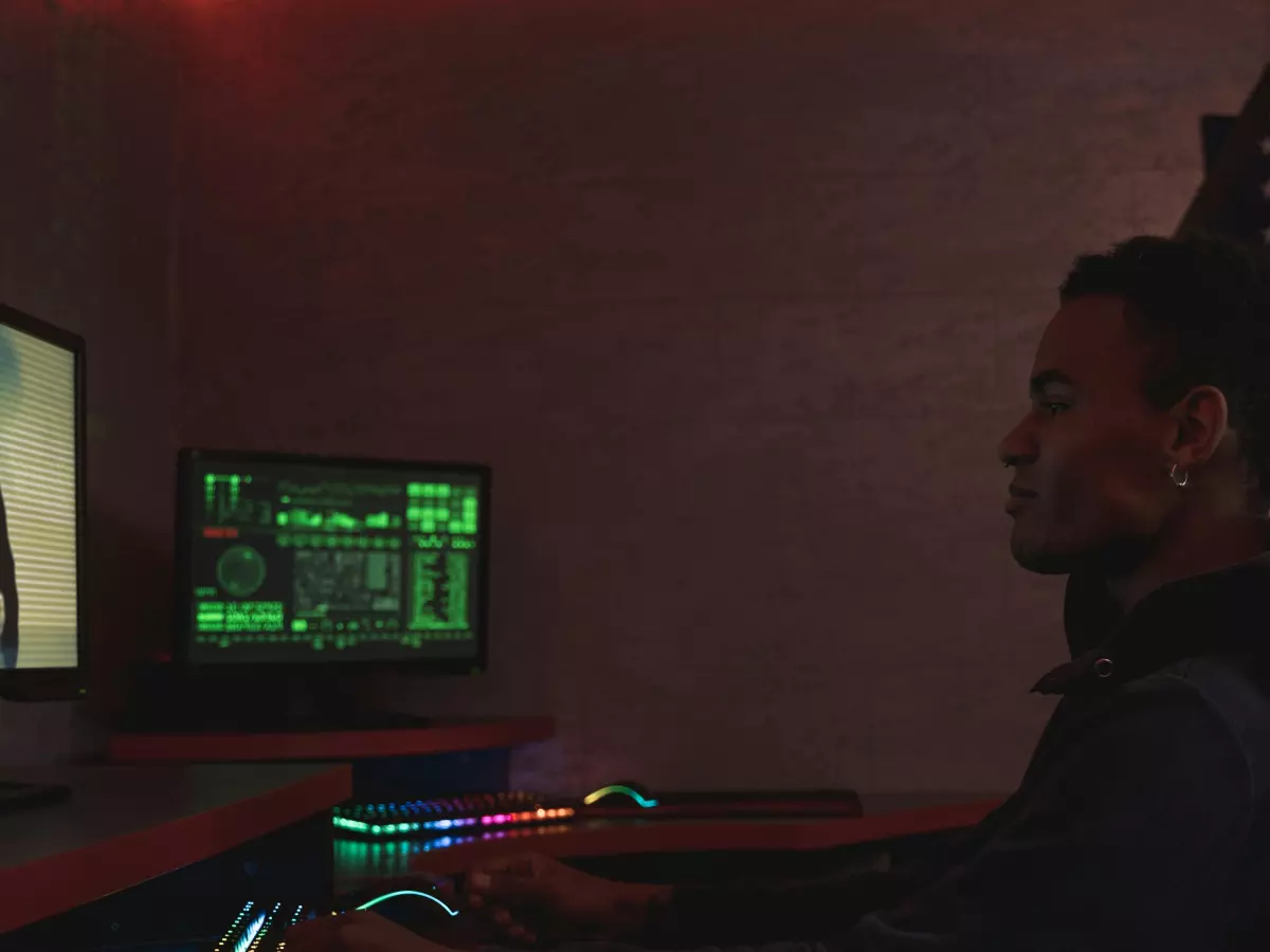 A man sits in a darkened room, hunched over a computer, his face illuminated by the screen. He is wearing a dark shirt and appears to be focused on the screen, which shows a close-up of lines of code. The room is dimly lit, with red lighting casting a sinister glow on the scene.