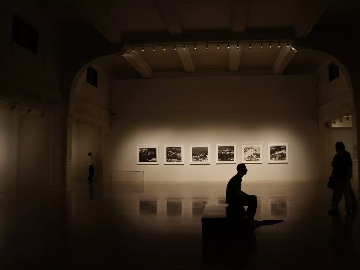 Silhouettes of people in a gallery. The focus is on a man sitting on a plinth while looking at a framed picture of the game artwork, while a woman is walking towards the right side of the image. The entire image is in dim lighting.