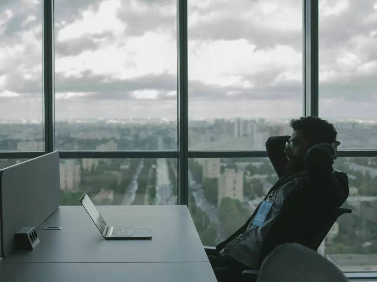 A person in a dark jacket sits at a desk in front of a large window with a city view, looking at a laptop. 