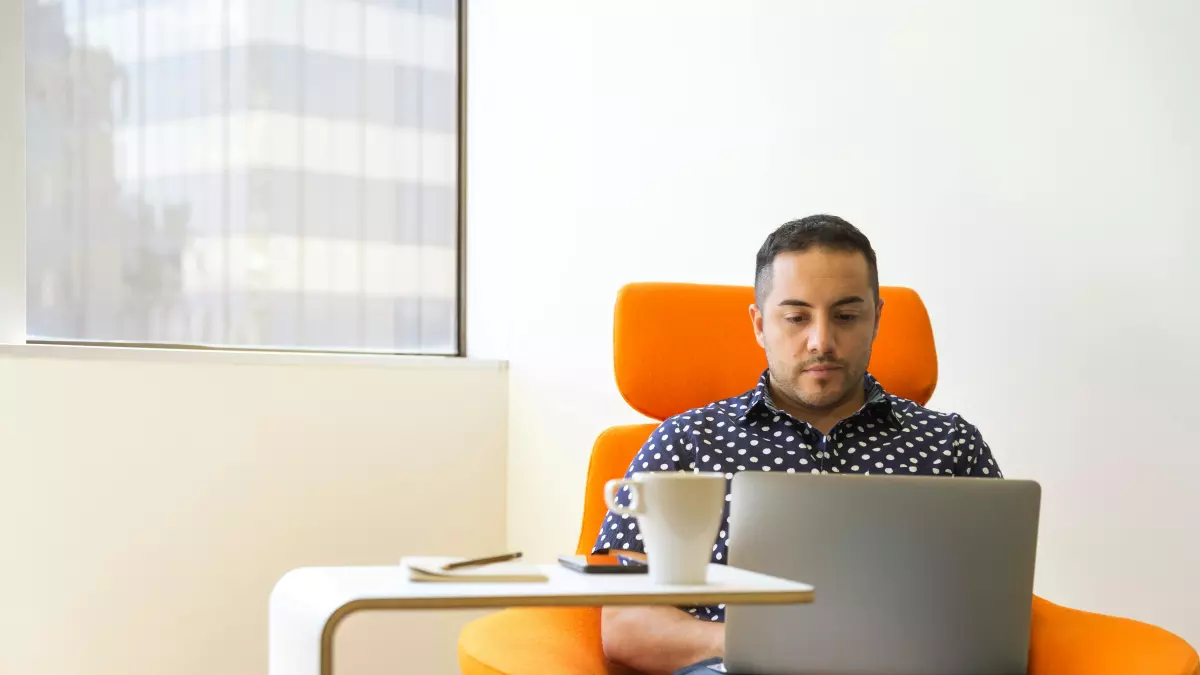 A man sitting in an orange armchair in front of a window, looking at his laptop. He