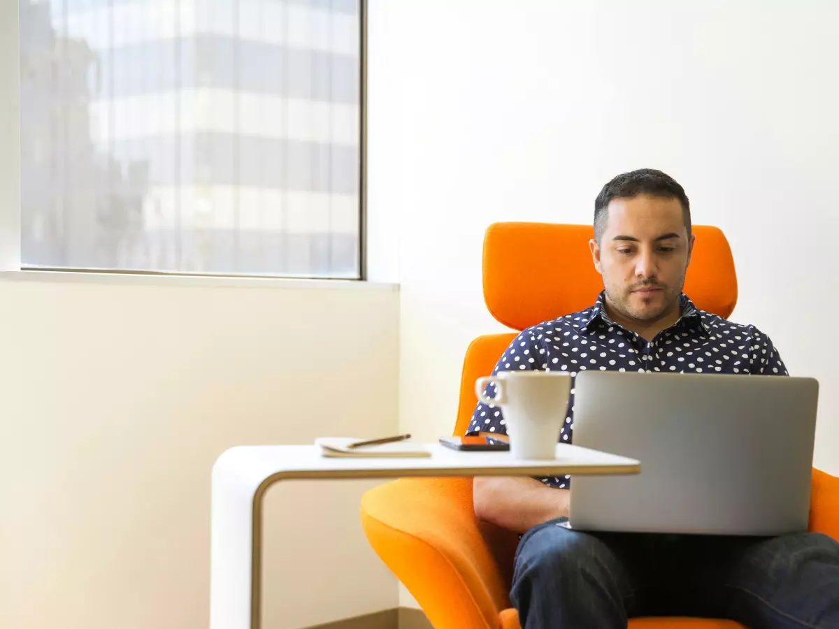 A man sitting in an orange armchair in front of a window, looking at his laptop. He's wearing a checkered shirt, jeans, and has a beard. He's holding a white coffee mug in his left hand. There is a small table in front of him with a smartphone and a pen on it.