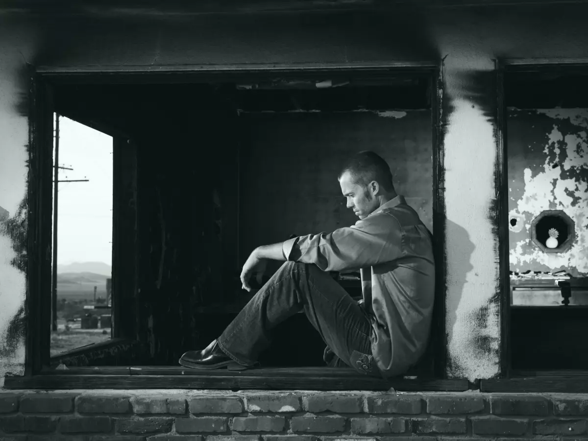 A man sits in a window, looking out. The image is in black and white, and the man's face is obscured.