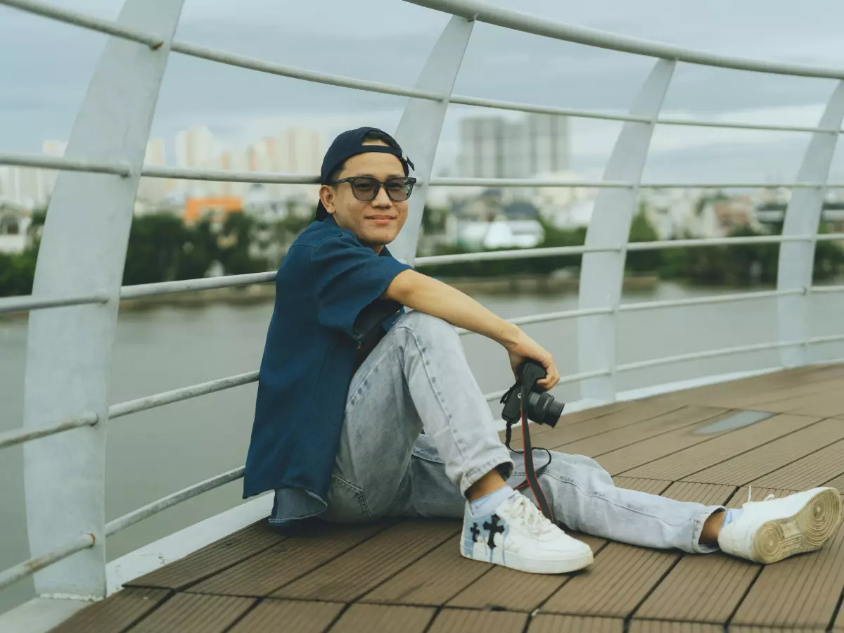 A young man sitting on a bridge, wearing a blue shirt and grey pants, he is holding a camera in his left hand, he is wearing a cap and sunglasses, he is looking at the camera