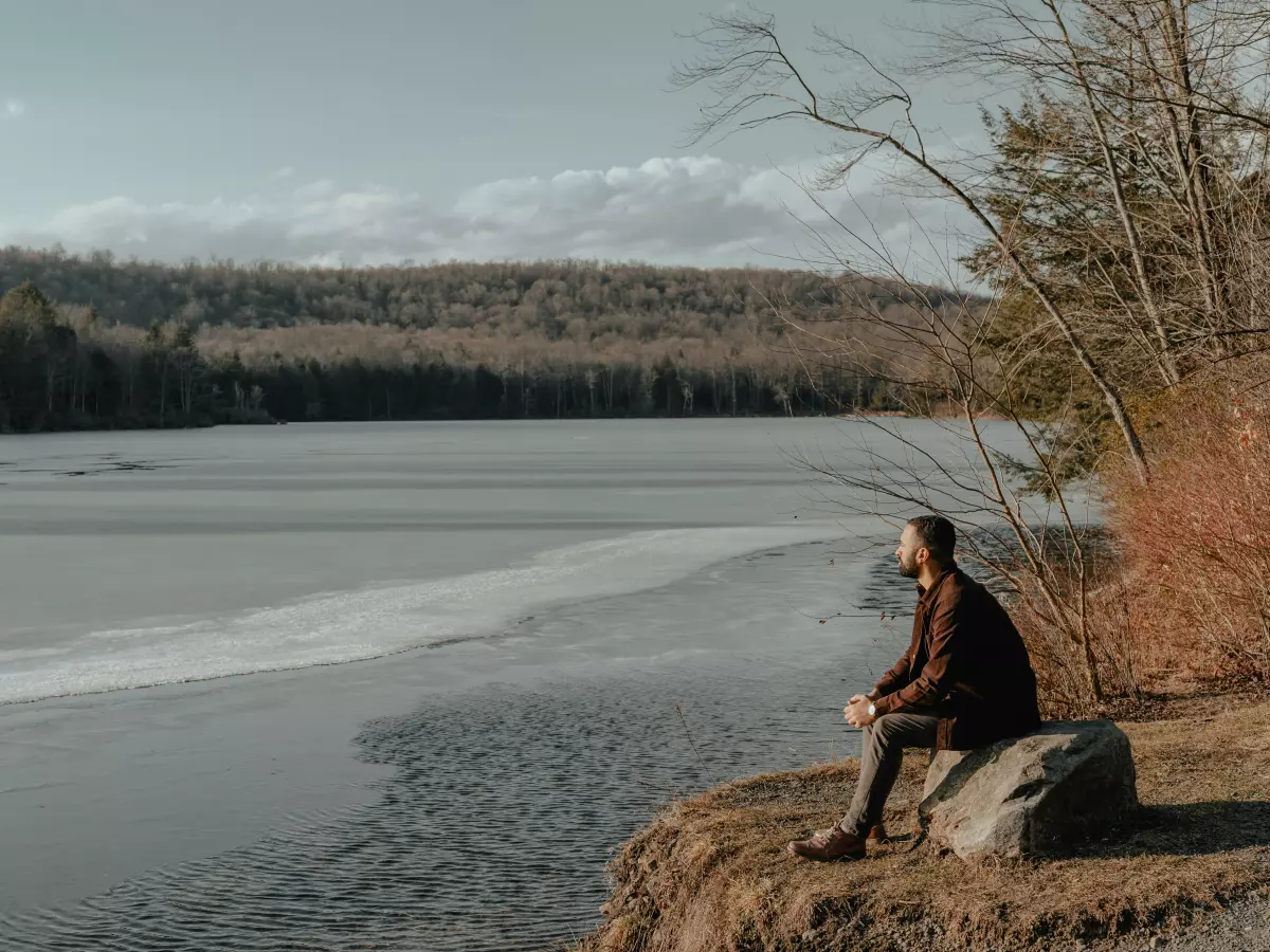 A man sits on a rock by a lake, looking out at the water. The lake is partially frozen, and there are trees in the background.