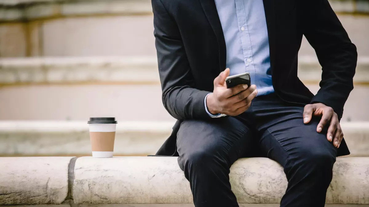 A person in a business suit is sitting on a stone wall and using a smartphone.