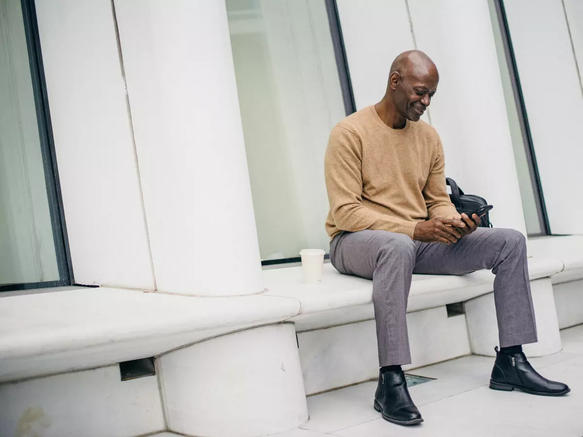 A man wearing a beige sweater and gray pants sits on a white ledge, looking down at his smartphone.