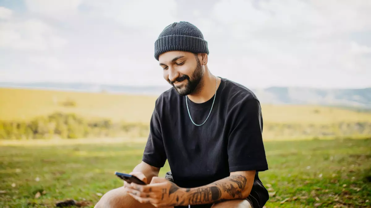 A man with a beard and a black beanie is sitting on a wooden bench in a field, looking at his phone with a smile on his face.