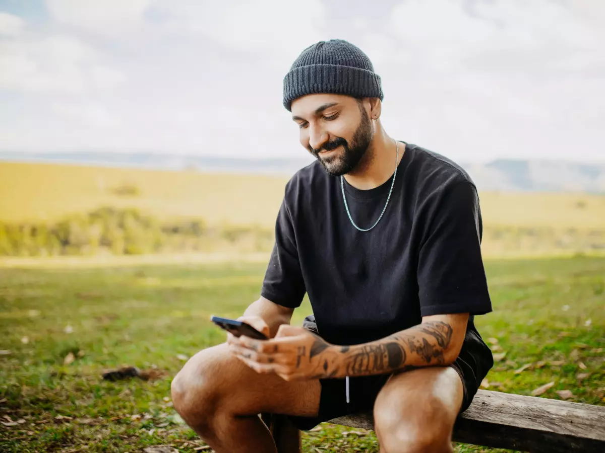 A man with a beard and a black beanie is sitting on a wooden bench in a field, looking at his phone with a smile on his face.