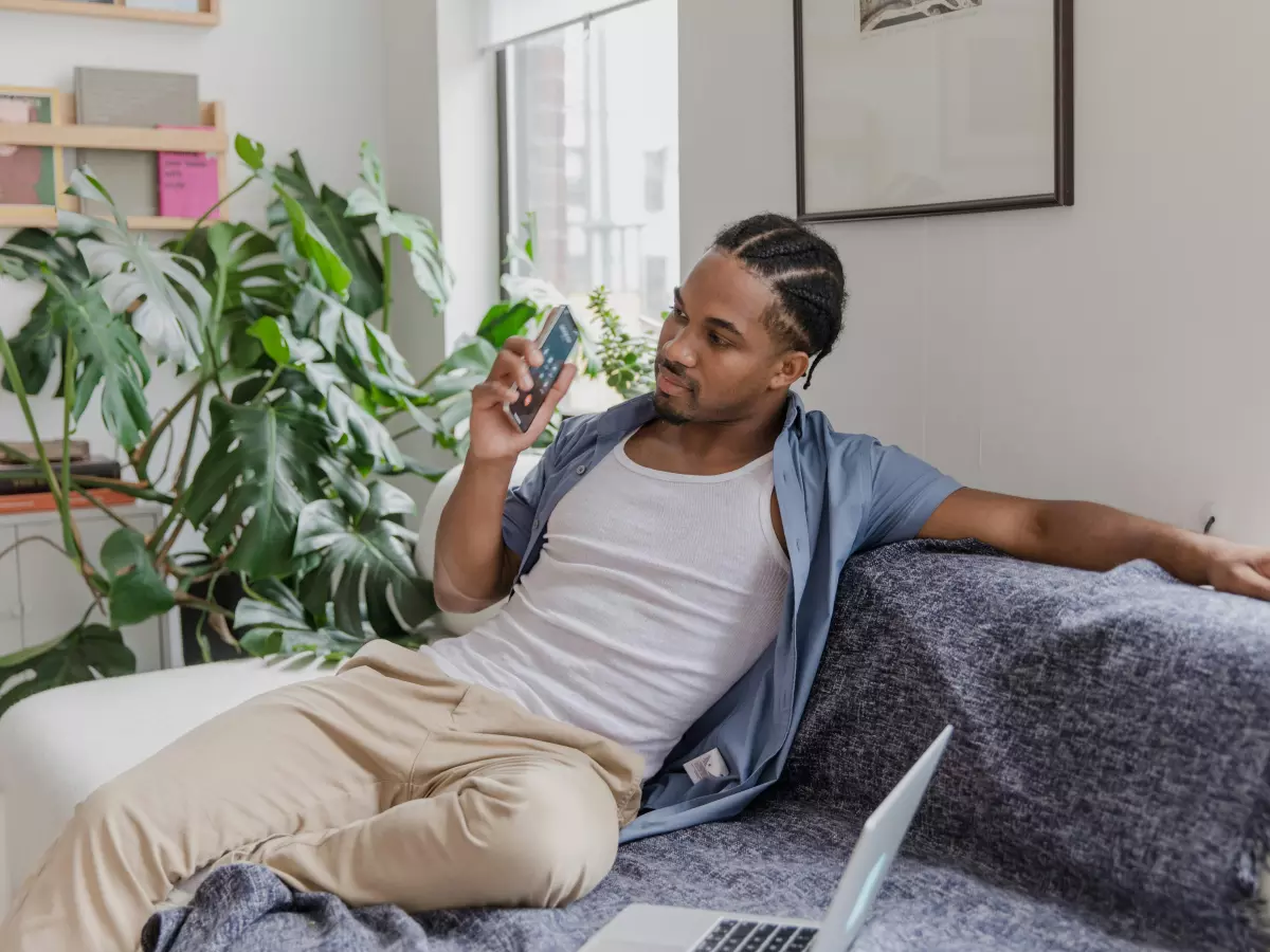 A young man is sitting on a couch in a living room, holding a smartphone and looking at it. He is wearing a white tank top and khaki pants. There is a potted plant in the background.