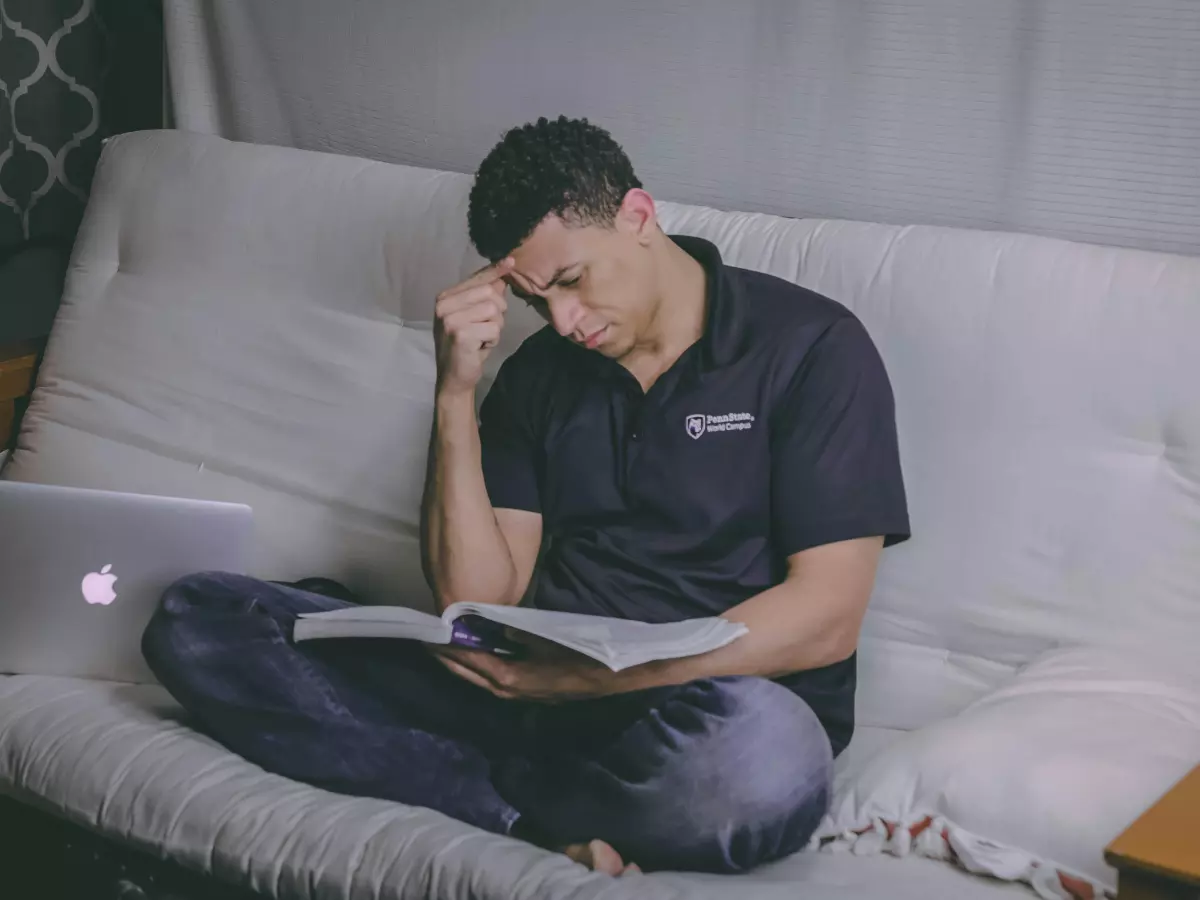 A man sitting on a couch, looking worried, reading a book. A laptop is visible on the couch behind him.