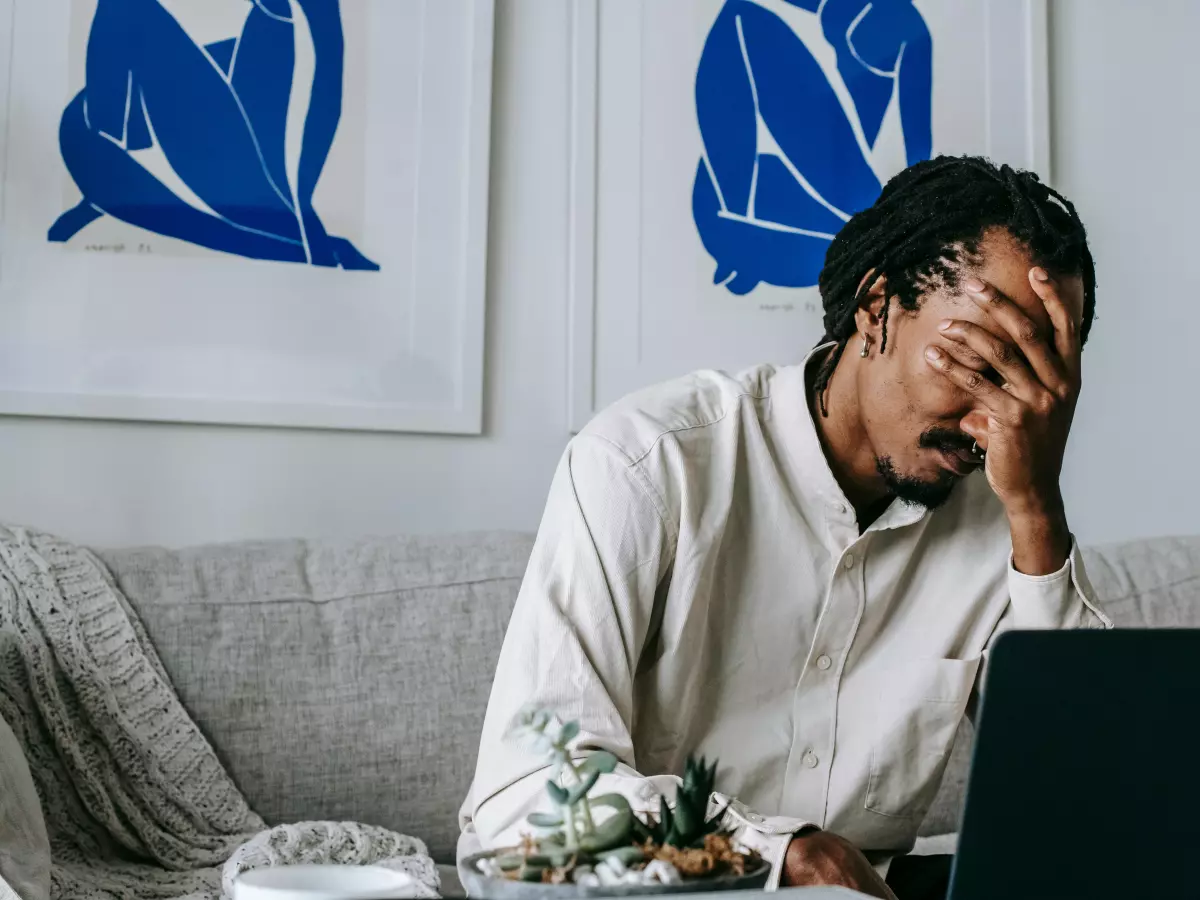 A man sits on a couch, covering his face with his hand, looking distressed. A laptop is in front of him, suggesting he's dealing with a problem related to technology. Money is scattered around, adding a financial dimension to the frustration.