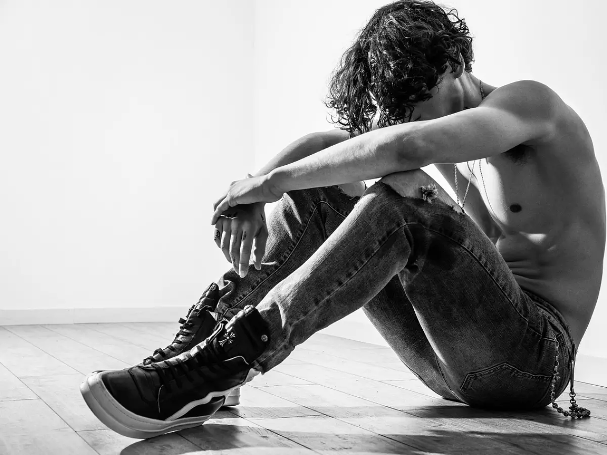 A young man is sitting on the floor with his head in his hands. He is dressed in jeans and a shirt and is looking down at the floor.