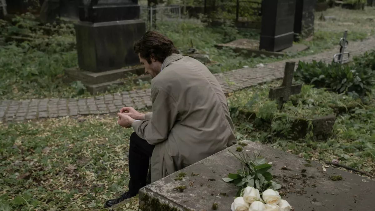 A lone figure is seated in a cemetery, with white flowers placed in front of a headstone.