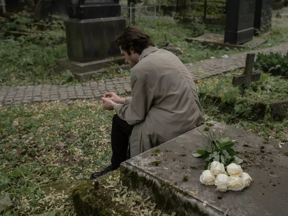 A lone figure is seated in a cemetery, with white flowers placed in front of a headstone.