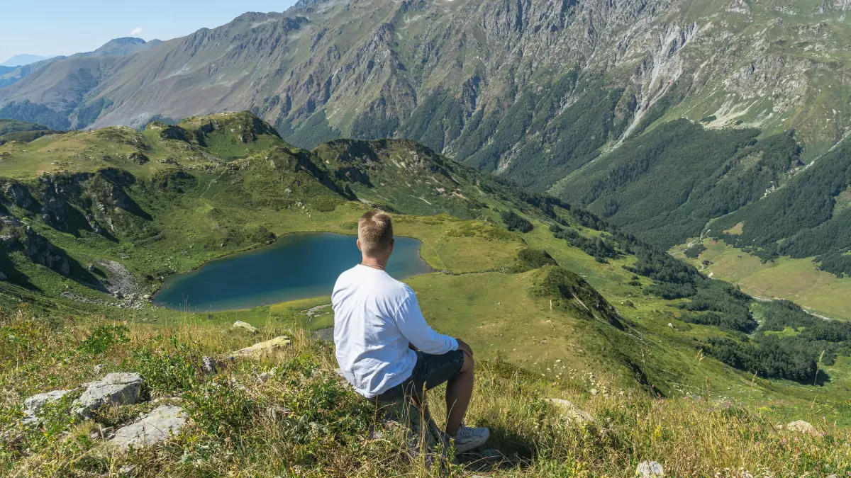 A person sits on a rock overlooking a valley with a lake. The person is wearing a white shirt and blue jeans. The sky is blue and the mountains are green.