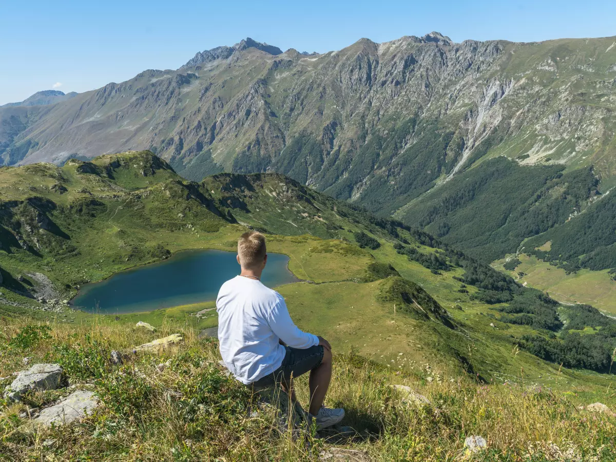 A person sits on a rock overlooking a valley with a lake. The person is wearing a white shirt and blue jeans. The sky is blue and the mountains are green.