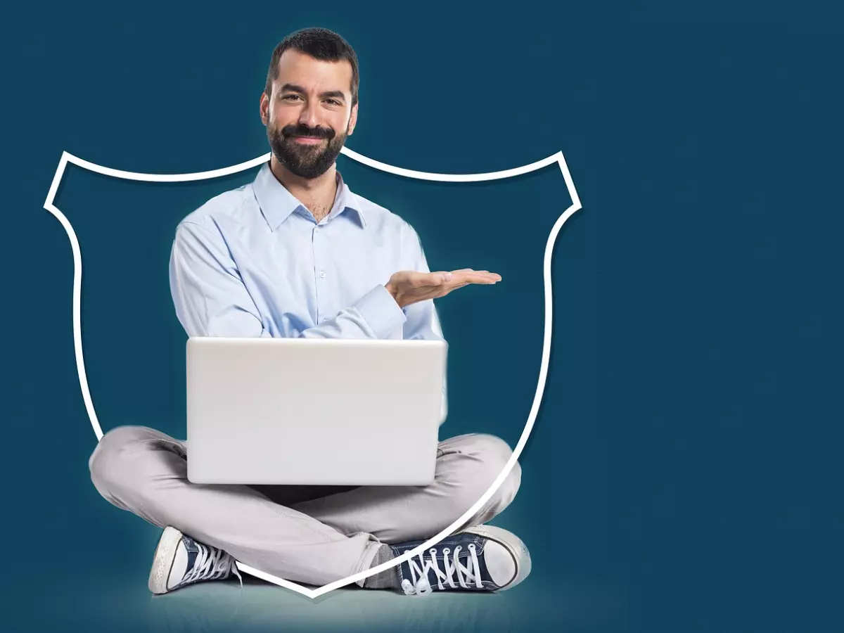 A man sits cross-legged on the floor, holding a laptop and smiling, a large shield icon behind him.