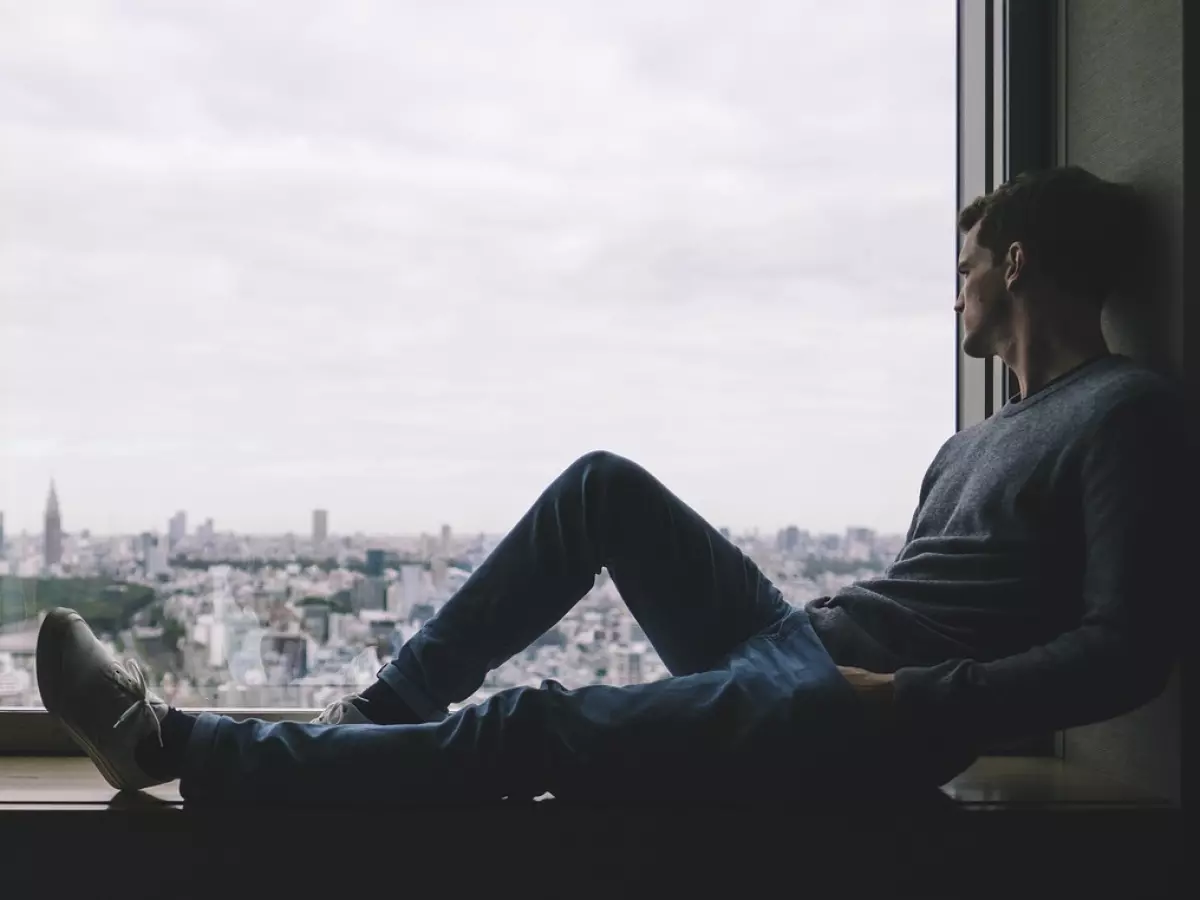 A man sits on a windowsill, looking out at a city skyline. He's wearing a sweater and jeans.