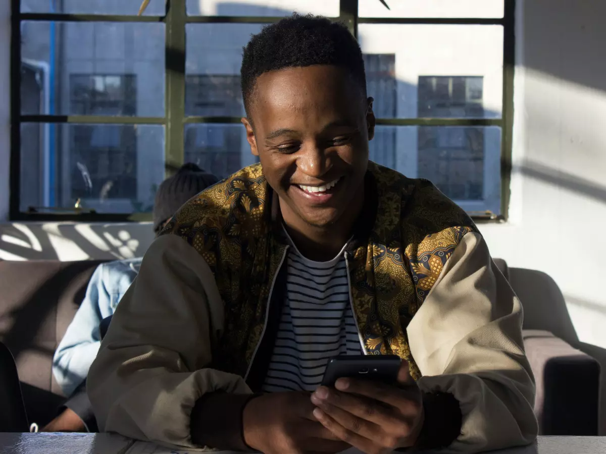 A young man in a patterned jacket sits on a couch and looks down at his phone. He is smiling. The lighting is bright and the room is spacious.