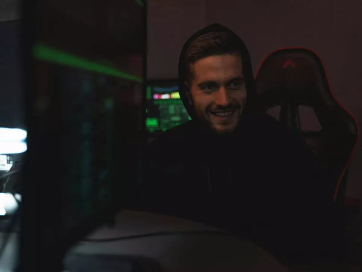 A man with a beard and headphones is sitting in front of a computer screen, looking intensely at the screen. The background is dark, highlighting the man and the screen. He is wearing a black hoodie and a black t-shirt.