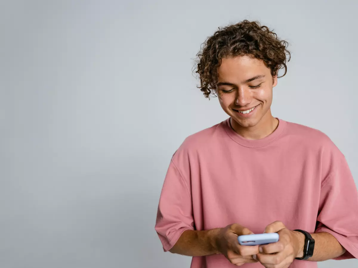 A young man with curly hair, wearing a pink t-shirt, smiles as he uses his smartphone, his face is illuminated by the screen.