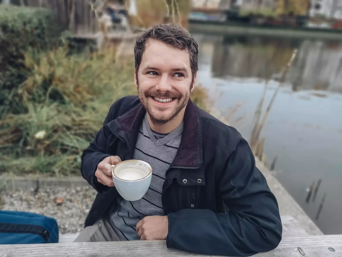 A man with a beard, sitting on a wooden bench by a pond.  He is looking off to the side, smiling and holding a coffee cup.