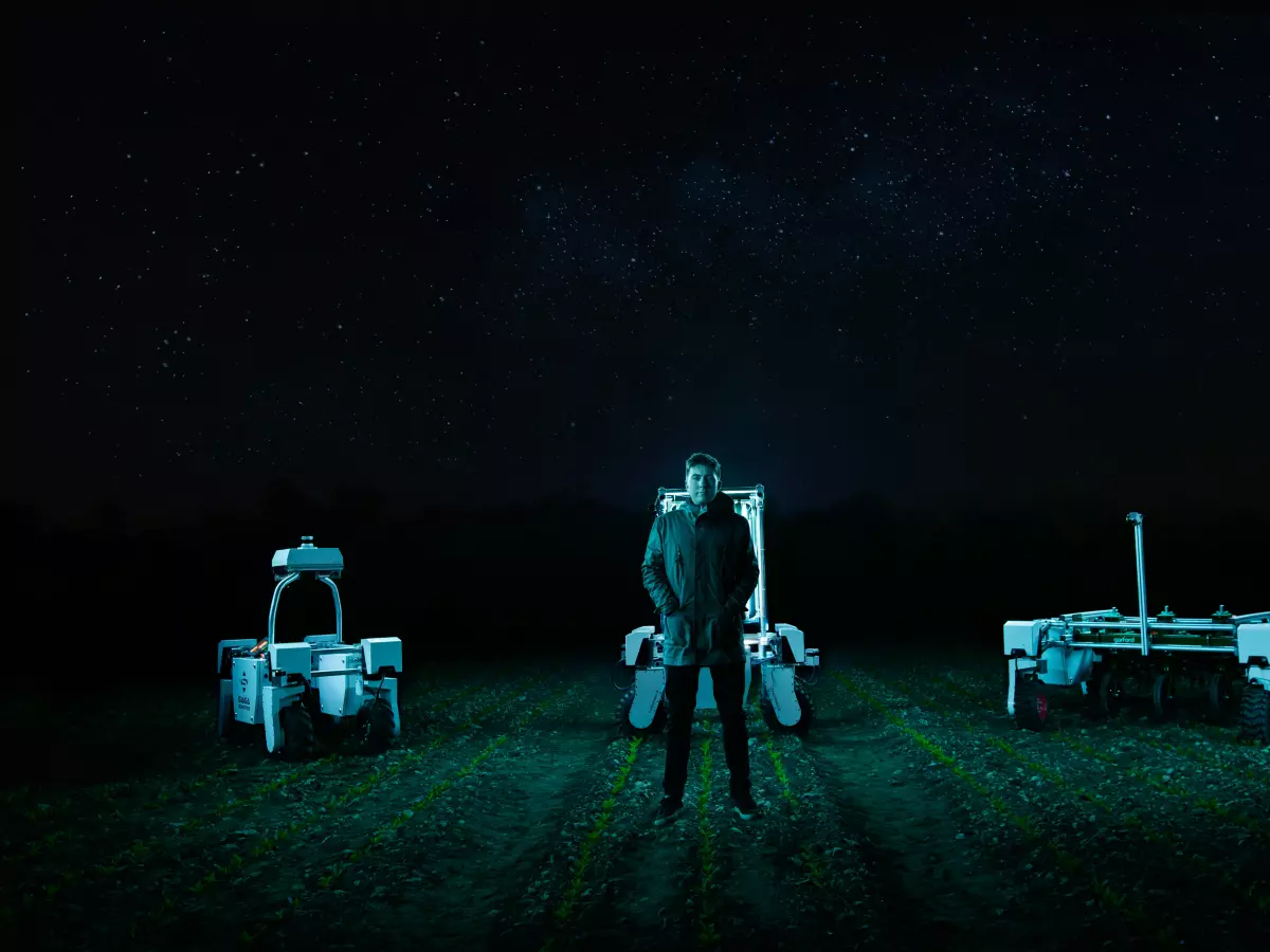 A man stands in the middle of a field at night, under a starry sky, with two autonomous robots on either side of him.