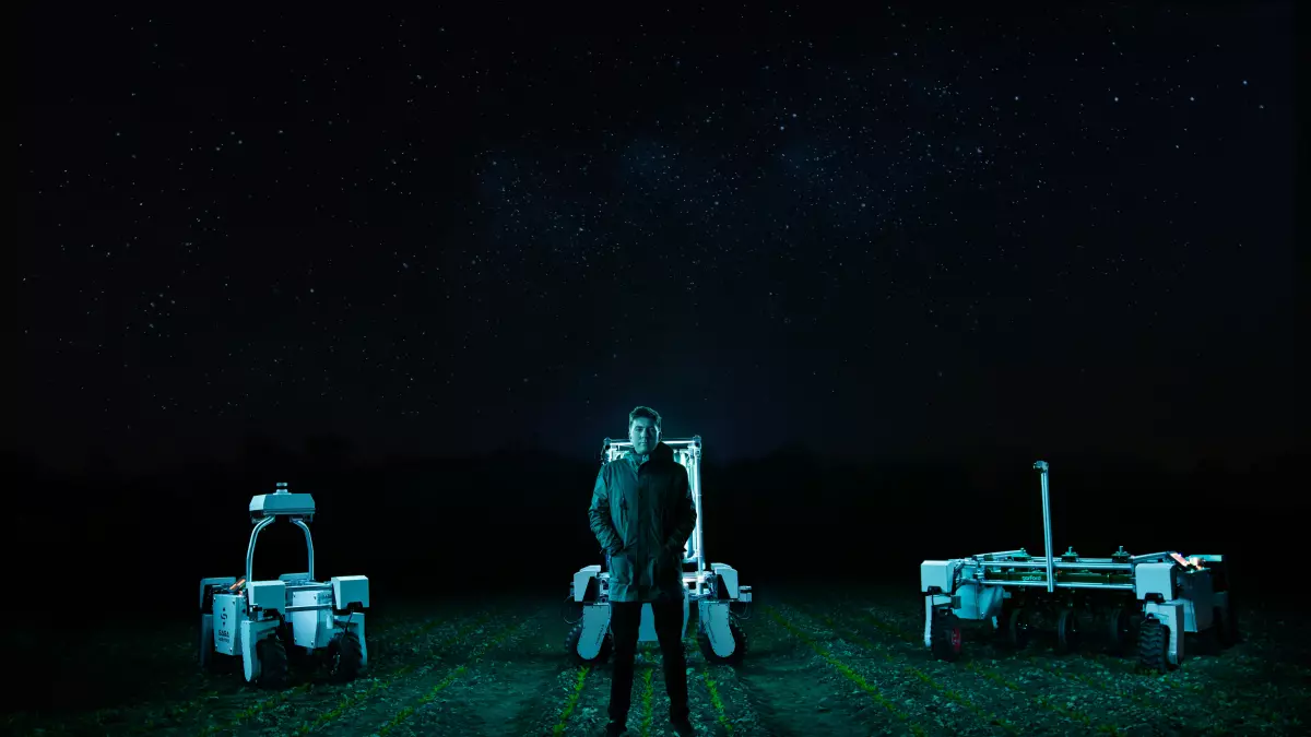 A man stands in the middle of a field at night, under a starry sky, with two autonomous robots on either side of him.