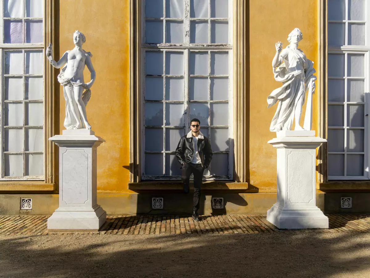 A person wearing a black coat is sitting on a bench in front of two white statues in a park.