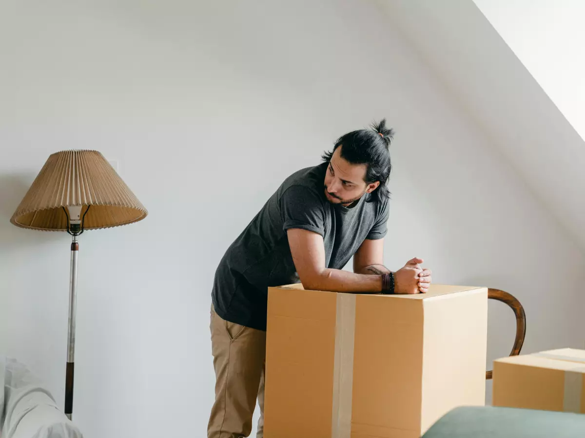 A man in a grey t-shirt and khaki pants leans against a cardboard box in a room with a lamp.