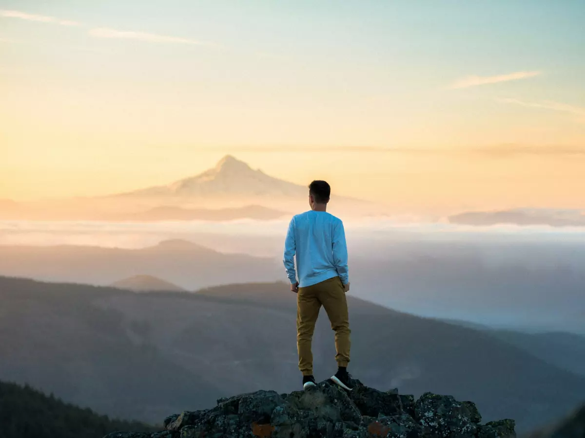 A person stands on a mountaintop, looking out at a vast expanse of clouds and mountains under a clear sky. The person is facing away from the viewer and is wearing a blue shirt and brown pants. The sky is a pale blue with hints of orange and pink from the sunrise.