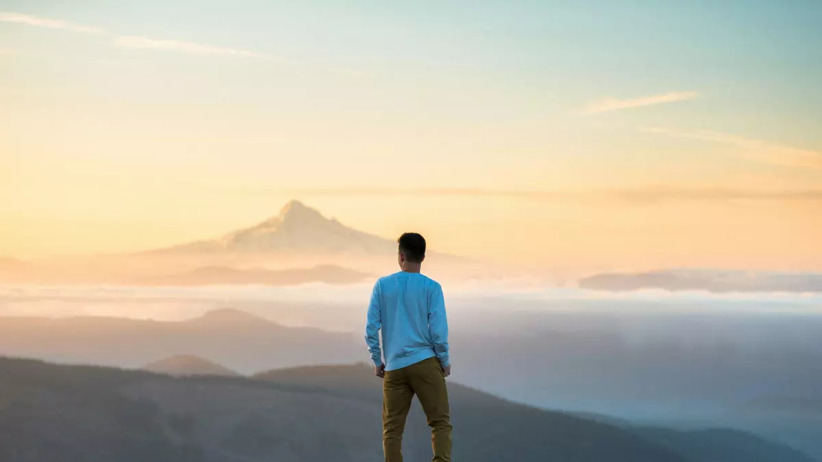 A person stands on a mountaintop, looking out at a vast expanse of clouds and mountains under a clear sky. The person is facing away from the viewer and is wearing a blue shirt and brown pants. The sky is a pale blue with hints of orange and pink from the sunrise.