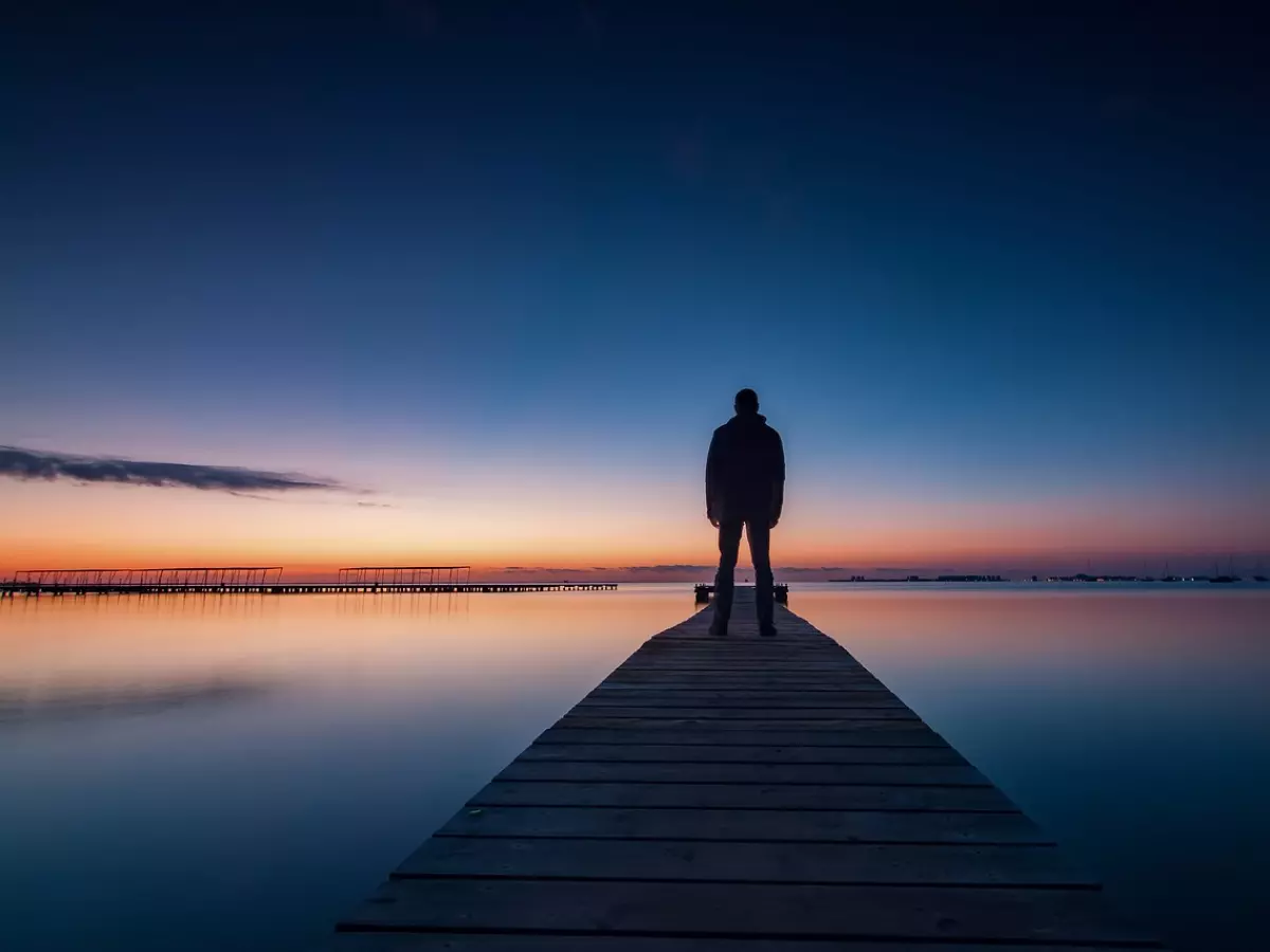 A person stands on a pier overlooking a body of water at sunset. The person is silhouetted against the sky, with their back to the viewer.