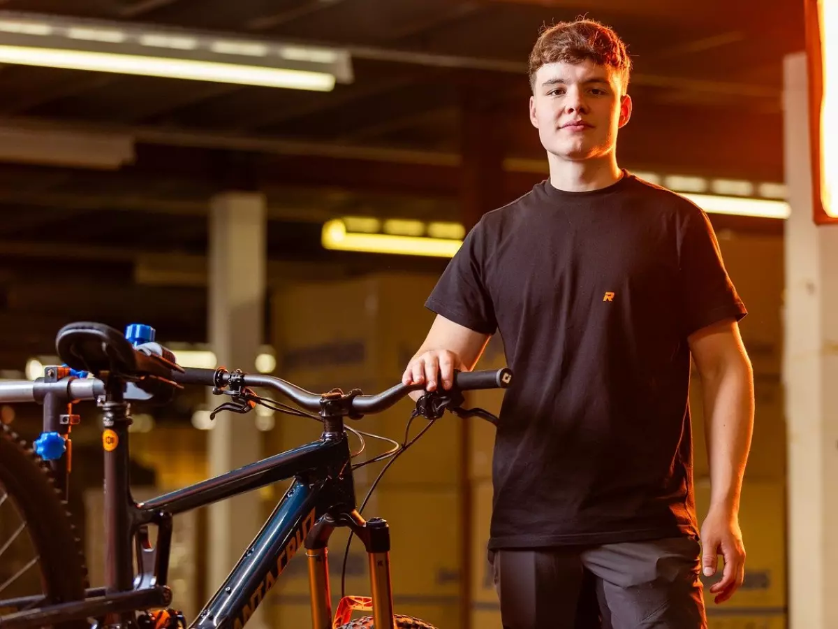 A young man in a black t-shirt stands next to a mountain bike in a garage setting.