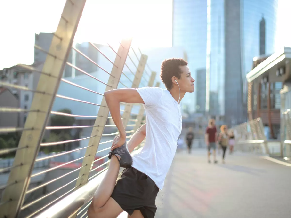 A man wearing wireless earbuds is stretching on a bridge in a city.