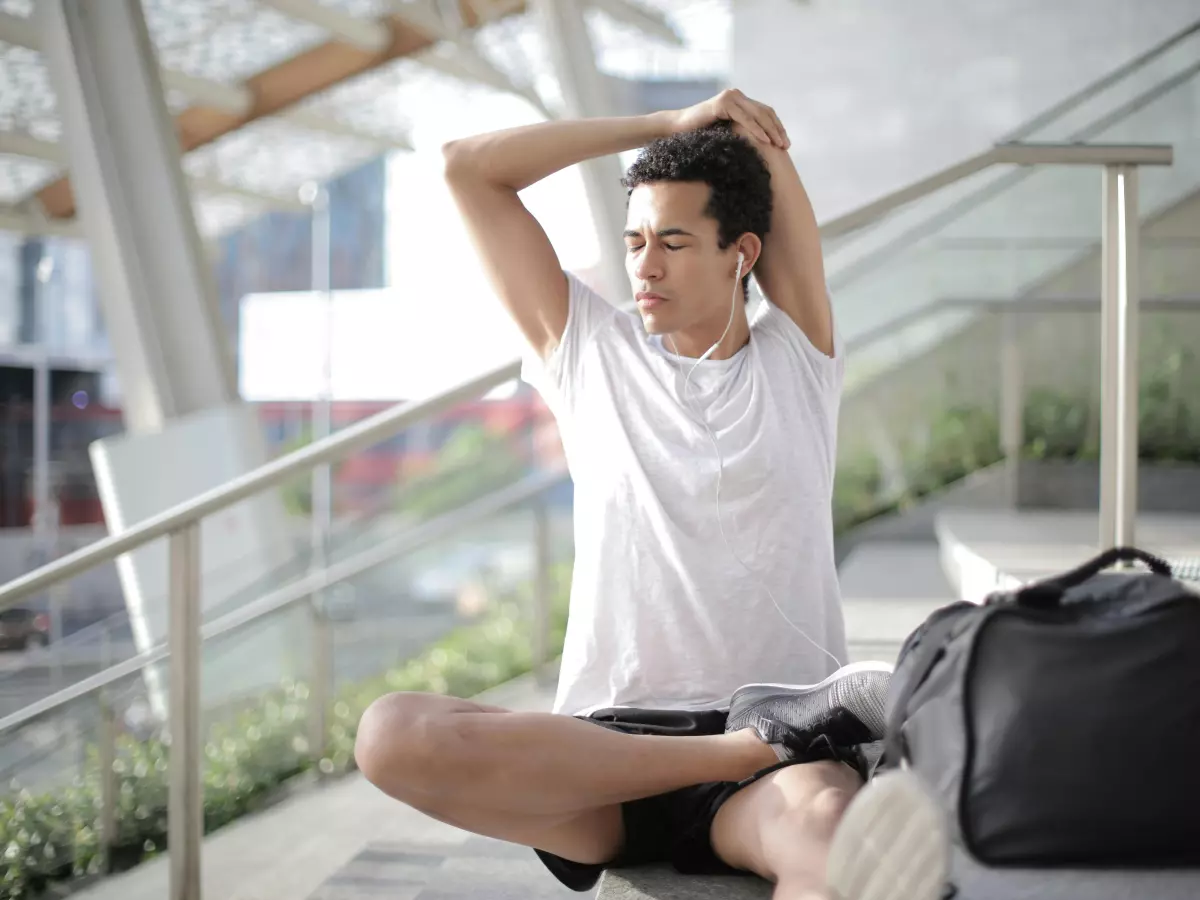 A young man wearing white t-shirt and black shorts is sitting on the stairs, hands behind his head stretching while wearing earphones. He has a black gym bag beside him. 