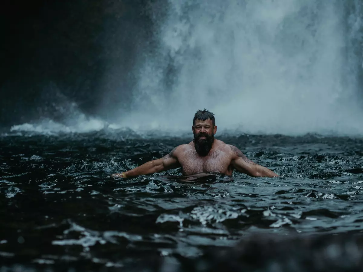 A man with a beard is standing in a river with a waterfall behind him. He is looking at the camera with a serious expression on his face. The water is rushing over the rocks and the scene is very dramatic.