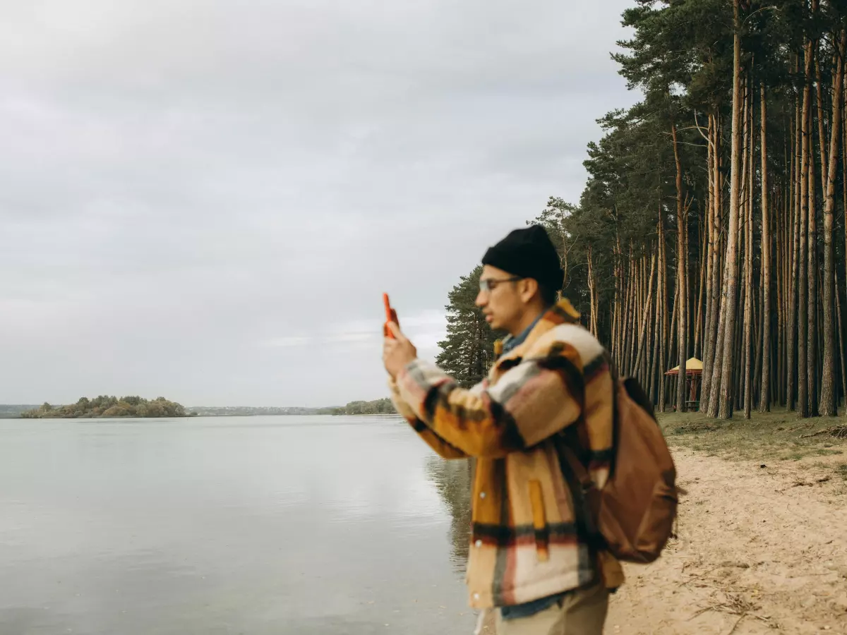 A man in a plaid jacket and black hat is standing on a lakeshore holding a smartphone in his hands. He is looking down at the phone. The background is a lake with pine trees and an overcast sky.